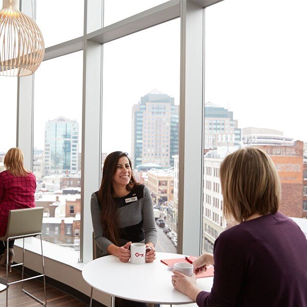 Two women sitting at a table holding coffee mugs, city landscape in the background