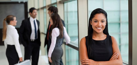Young woman with her arms crossed in front of a corporate office window with men in suits talking in the background