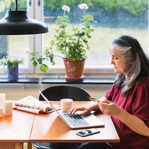 An older woman sits at a laptop at a kitchen table and enters her credit card number