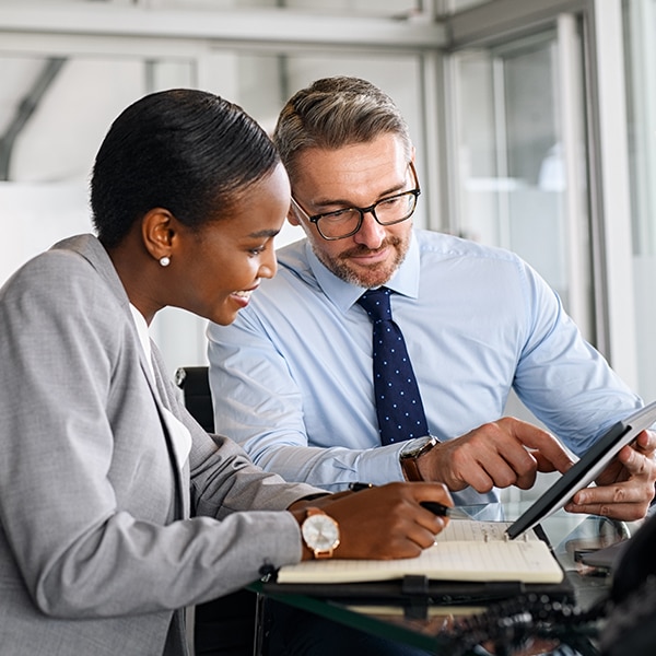 Mature businessman discussing work with black manager writing notes in diary. Confident business man in formal clothing working with african american woman showing data on digital tablet. Smiling professional business people working together on new business strategy.
