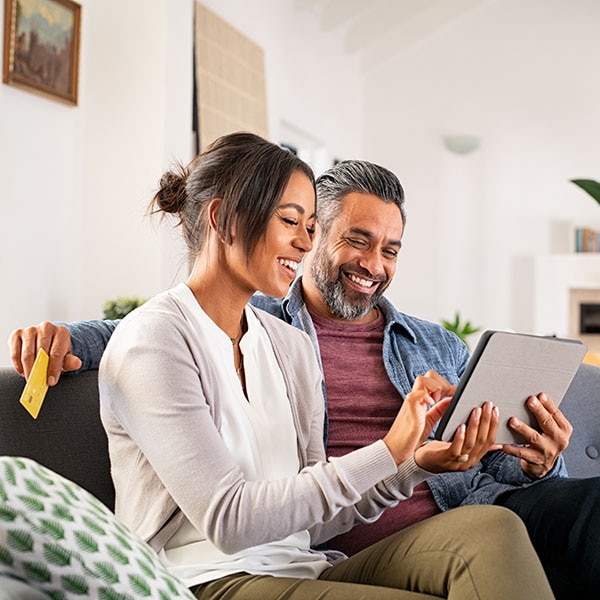 Happy multiethnic mature couple using digital tablet for online payment with credit card. Cheerful latin wife showing something to buy on digital tablet to her indian husband while holding bank card. Mid adult hispanic woman and middle eastern man sitting on couch in living room while doing shopping online together.