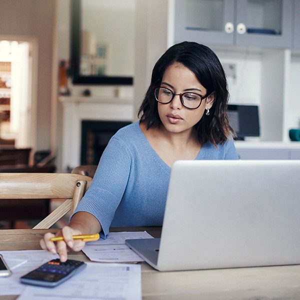 A woman working at a table at home, surrounded by paper and using both a laptop and a calculator