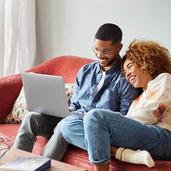 Two people sitting on a couch using a laptop together at home, reviewing information online in a relaxed living room setting.