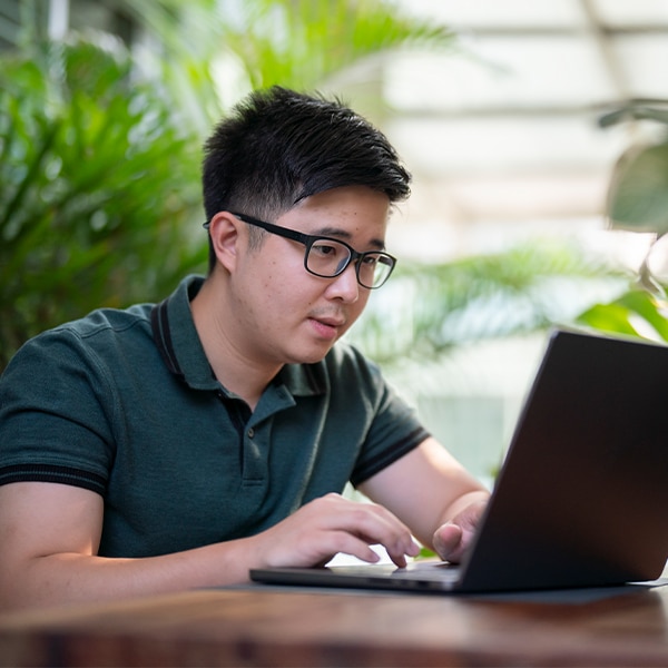Person working on a laptop at a table surrounded by plants, researching information online in a bright indoor workspace.
