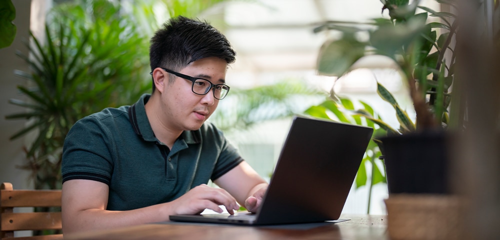 Person working on a laptop at a table surrounded by plants, researching information online in a bright indoor workspace.