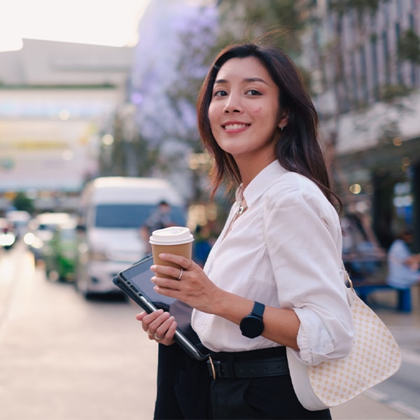 Person holding a takeaway coffee and tablet while standing on a city street.