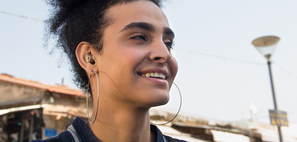Person outdoors wearing earbuds and hoop earrings, with buildings visible in the background.