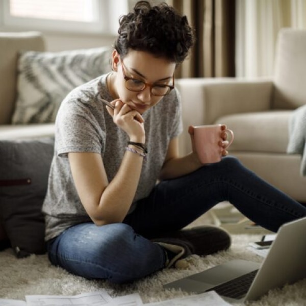 Person sitting on a living room floor reviewing student loan paperwork on a laptop, holding a coffee mug while managing finances at home.