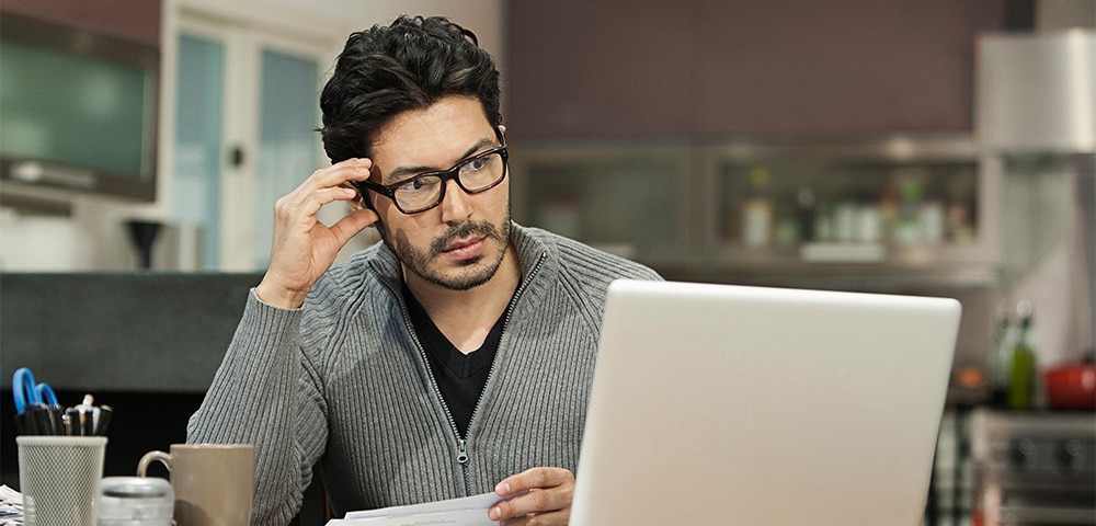 Person sitting at a table with a laptop and papers, appearing to review documents at home.